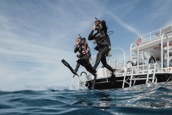 Two PADI Master Scuba Diver Trainers in wetsuits and gear jump off a boat into the ocean. The water is calm with a hint of waves, and the sky is partially cloudy. The boat is white, with ladders and equipment visible on deck.